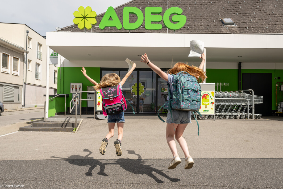 Two children with backpacks joyfully jump in front of a supermarket entrance on a sunny day.