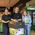 Three people smiling and standing inside a grocery store, with two of them holding cardboard boxes. There are fresh produce and refrigerated beverage displays in the background.