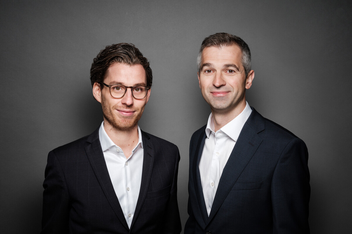 Two men in business suits standing against a dark gray background. Both are smiling slightly and looking directly at the camera, with the man on the left wearing glasses.