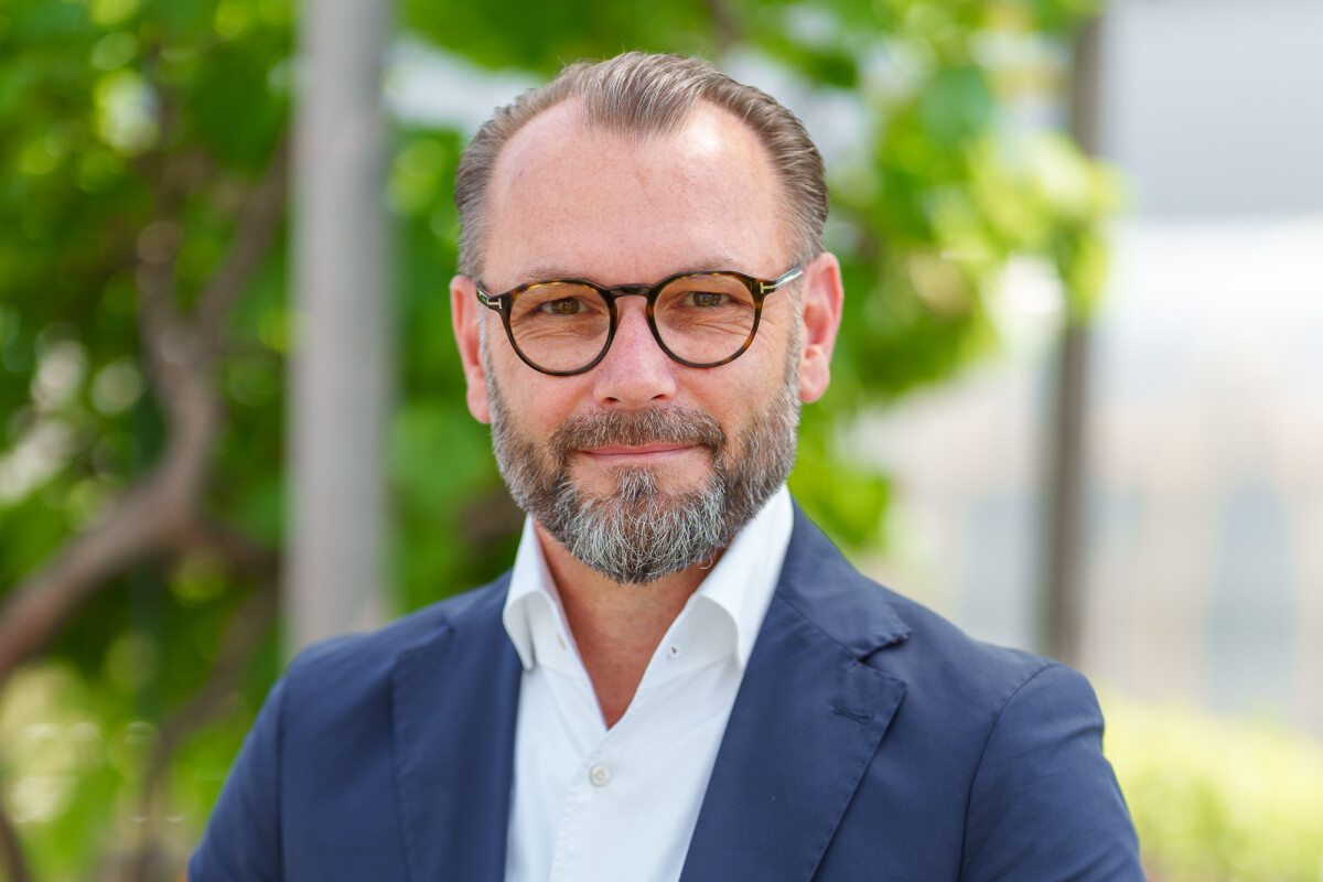 A man with a beard and glasses is wearing a navy suit and white shirt, standing outdoors with greenery in the background.