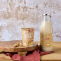 A rustic wooden table displays a cup labeled "Bio Hafer Natur" placed next to a glass bottle of oat milk. A small wooden slab holds a decorative sprig of oats, with a red cloth partially underneath. A textured wall serves as the background.