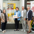 Five people standing together in front of a shop window and a promotional banner, smiling at the camera. One person holds a sign with the text "Genussland Oberösterreich." The group appears to be in a city setting.