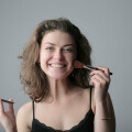 A smiling woman with curly brown hair applies makeup with a brush while holding a compact case. She is wearing a black top against a gray background.