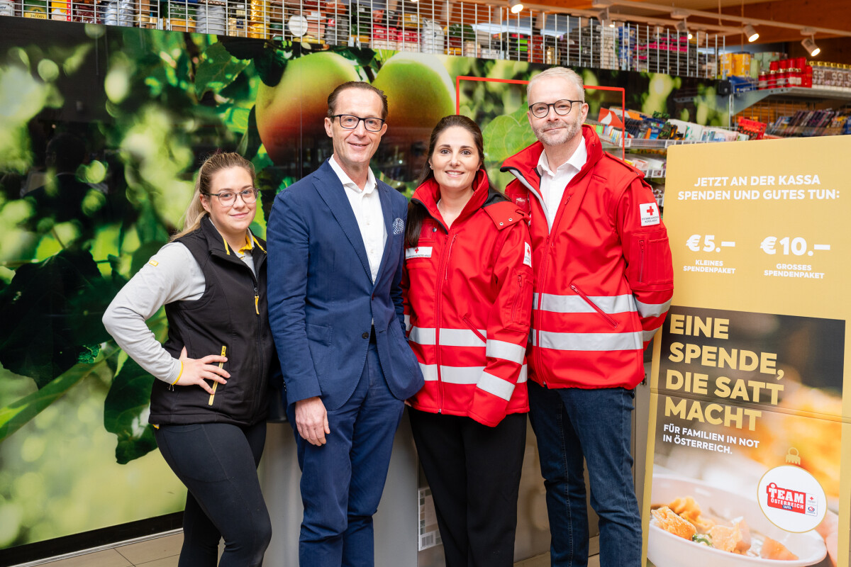 Four people stand together in a store, with two wearing red jackets with reflective stripes. They are next to a donation poster. The background features shelves and a large fruit-themed mural.