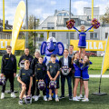 A group of people, including a person in a purple elephant mascot costume, football players, and cheerleaders, stand on a football field. Two large yellow flags with the "BILLA" logo are displayed, and a cheerleader holds pom-poms while being lifted in the air.
