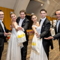 A group of three men and two women dressed in formal attire, with the women wearing tiaras and holding white bags. They are smiling and posing together on a wooden floor in a hall with high ceilings.