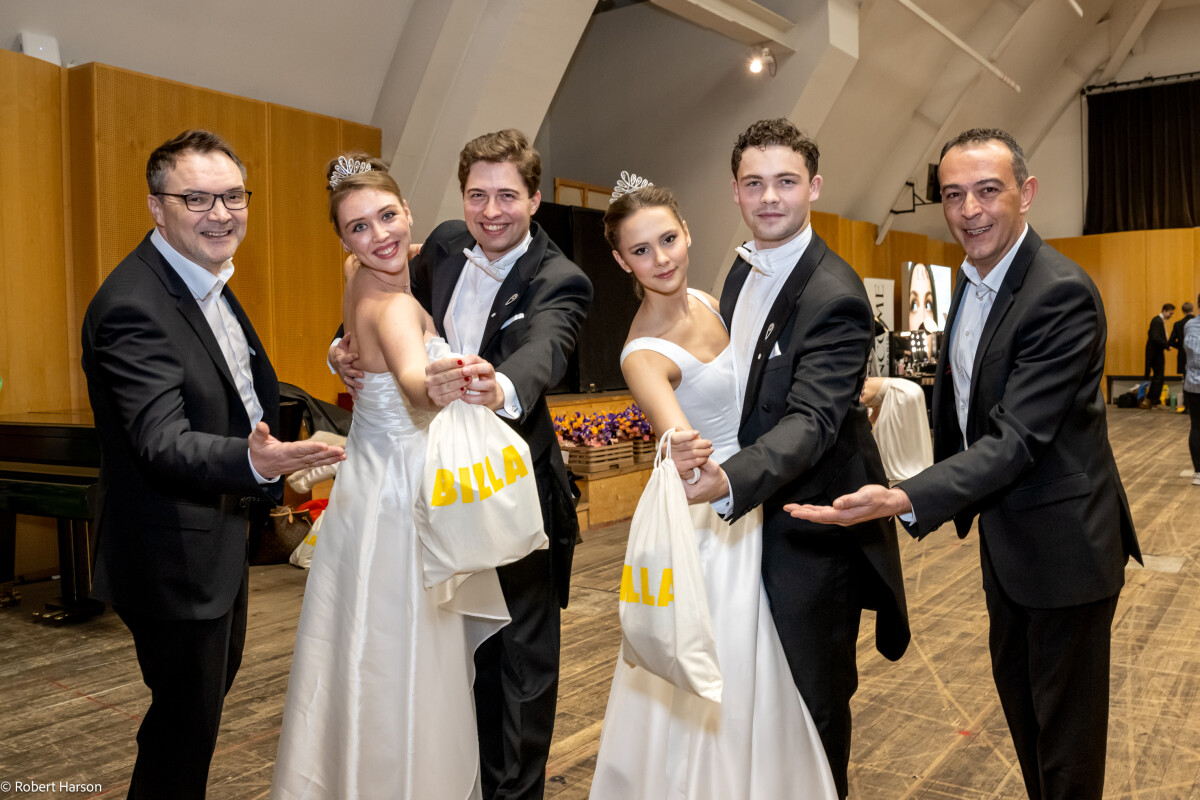 A group of three men and two women dressed in formal attire, with the women wearing tiaras and holding white bags. They are smiling and posing together on a wooden floor in a hall with high ceilings.