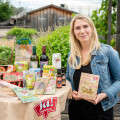 A woman standing next to a table covered with various food products. She is holding a package and smiling. The table displays items like bread, wine, and packaged goods. The setting appears to be outdoors with greenery in the background.