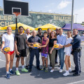 A group of people, including several wearing basketball jerseys, stand together on an outdoor basketball court. They are holding basketballs and posing for a photo. In the background, there are yellow tents and a large billboard. Two police officers are part of the group, and others are casually dressed. The scene appears to be a community event on a sunny day.