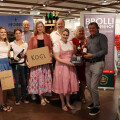 A group of smiling people standing together in a store, holding different wine bottles and boxes with various winery brands displayed. Some are dressed in traditional attire, and there are wine bottles on shelves in the background. Banners with winery names are visible.