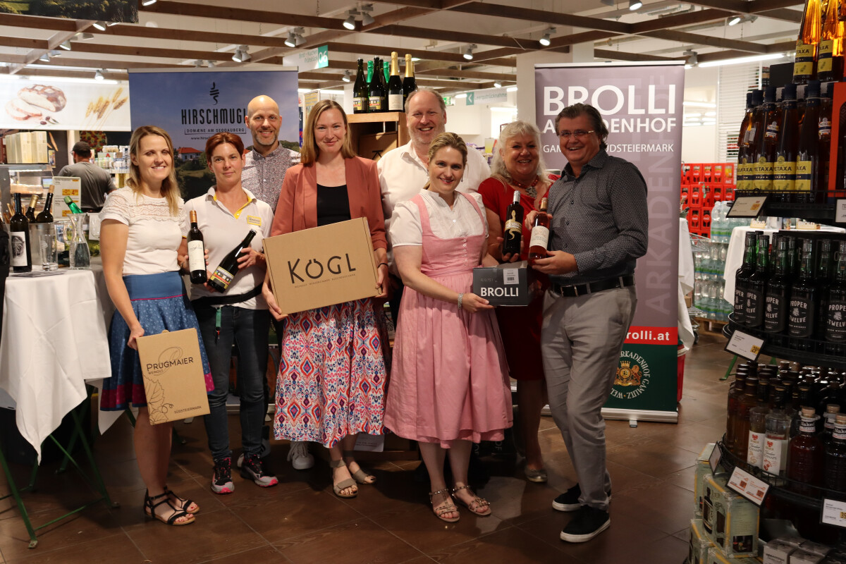 A group of smiling people standing together in a store, holding different wine bottles and boxes with various winery brands displayed. Some are dressed in traditional attire, and there are wine bottles on shelves in the background. Banners with winery names are visible.