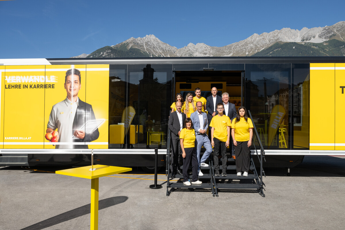 A group of people poses in front of a large yellow mobile unit with a mountainous backdrop. The unit features an advertisement with a person holding a tablet. Most individuals are wearing yellow shirts. A table with the same yellow stands to the side. The sky is clear and blue.