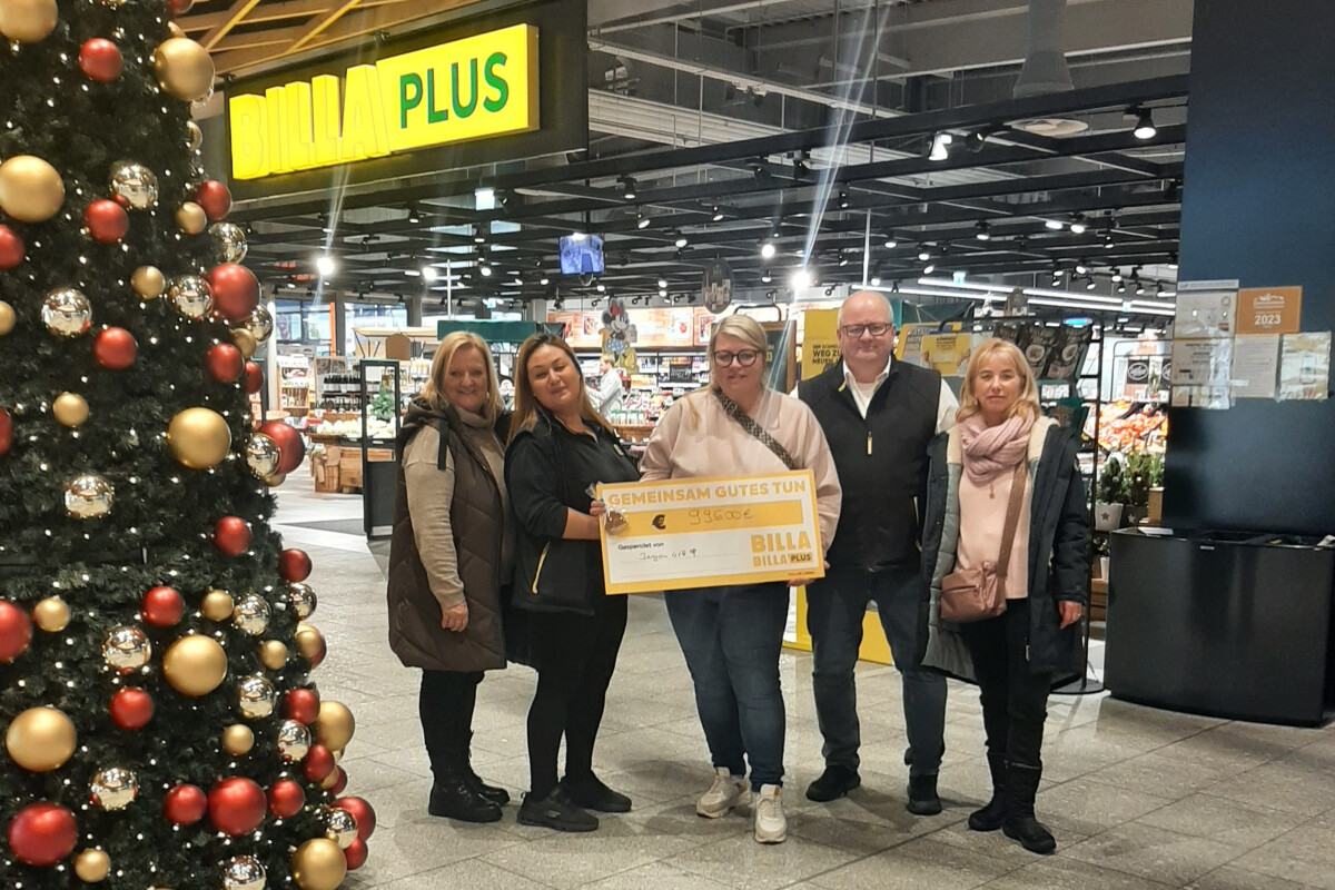 Five people pose together holding a large check in a supermarket near a decorated Christmas tree. The store's sign above reads "BILLA PLUS."