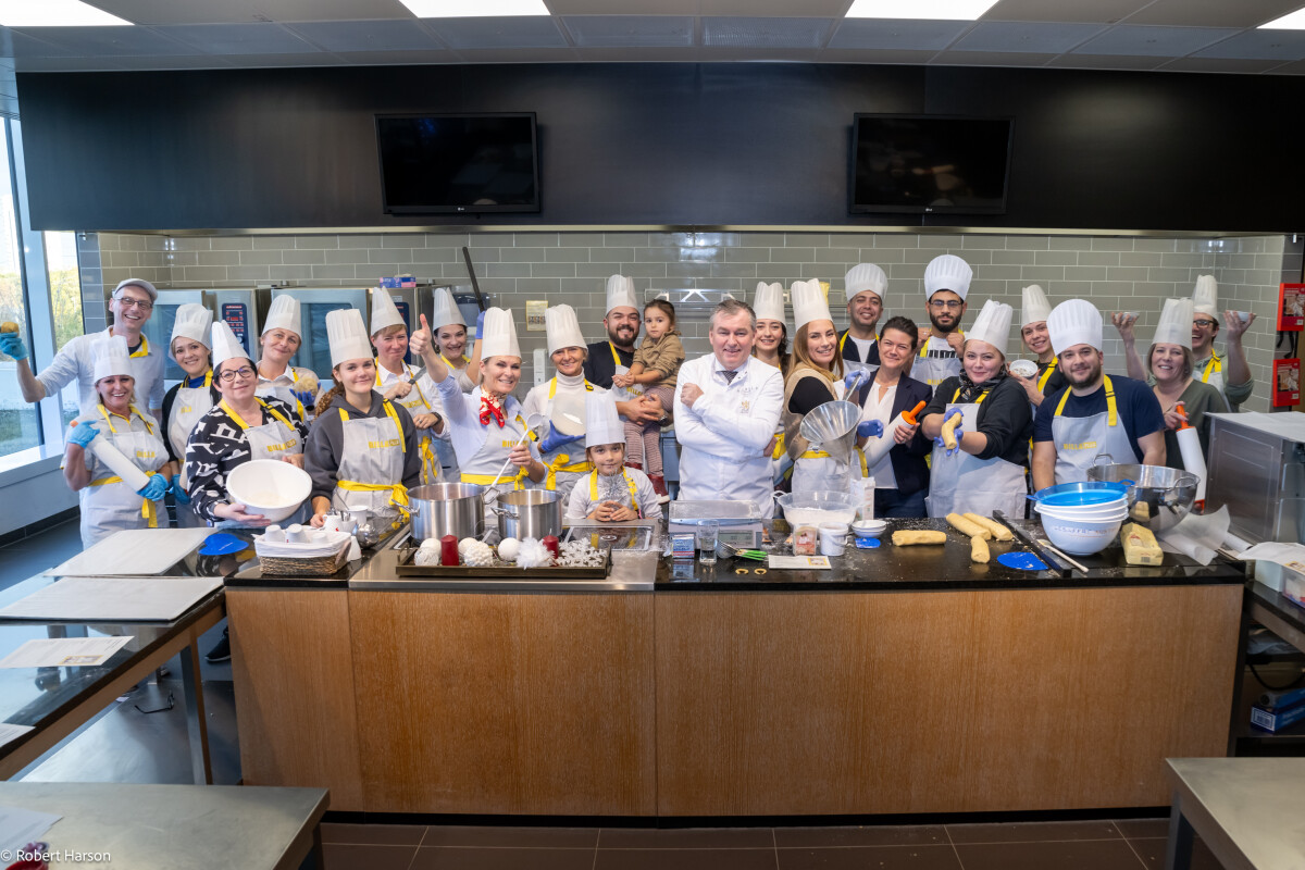 A group of people wearing chefs' hats and aprons stand smiling in a professional kitchen with cooking utensils and ingredients on the counter.