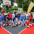 A diverse group of young basketball players and adults are gathered on an outdoor basketball court. The players are in blue uniforms, and several adults are standing behind them. Two large yellow banners with the word "BILLA" and a basketball hoop are in the background. Some of the kids are holding basketballs.