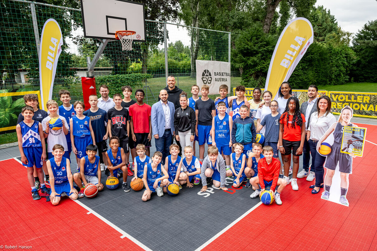 A diverse group of young basketball players and adults are gathered on an outdoor basketball court. The players are in blue uniforms, and several adults are standing behind them. Two large yellow banners with the word "BILLA" and a basketball hoop are in the background. Some of the kids are holding basketballs.