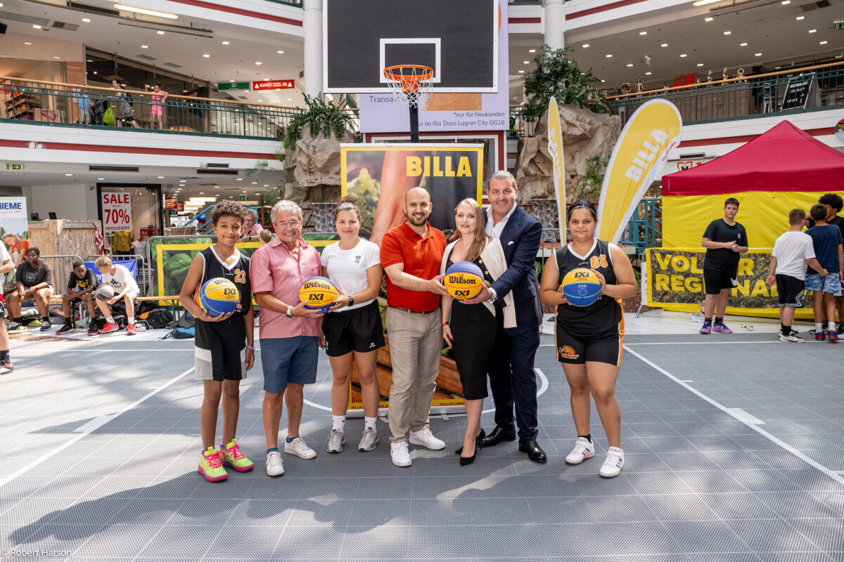 Seven people standing on a basketball court in a shopping mall, some holding basketballs. A banner and a basketball hoop are visible in the background, and spectators watch from the upper level.