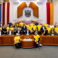 A diverse group of people is gathered in a formal room, with some wearing bright yellow shirts. They are positioned around a large wooden desk, and European and red-striped flags are displayed in the background. A large coat of arms hangs on the wall. The setting appears to be a government or council chamber.