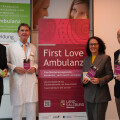 Four people stand in front of a display for "First Love Ambulanz" at a university clinic, holding informational brochures. The display has a pink and red design, and the clinic's details are shown on a board to the left.