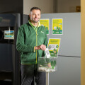 A man in a green jacket is smiling and standing in a room next to a machine, holding a clear box filled with small items. Posters with text and images are visible on the wall behind him.