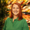 A smiling woman with red hair wearing a green collared shirt stands in front of a decorated Christmas tree. The background is softly illuminated with colorful lights.