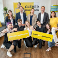 A group of people smiling in an office setting. Some are holding signs that say "#NEXTBILLAGENERATION". In the background, there is a yellow and white banner with text, and a TV screen displaying similar branding.