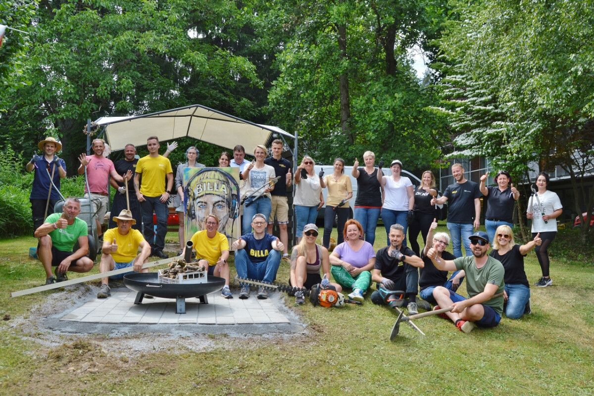A diverse group of people posing outdoors in front of a covered display featuring a large painted face. They are smiling and holding various gardening tools, with trees and greenery in the background. The group appears to be celebrating or completing a project.