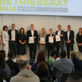A group of people standing in a row on a stage, holding awards. They are dressed in formal attire. An audience is seated facing them. A sign above reads "Betriebsrat Billa Wir Für Dich."