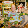 A person smiling and holding several Asterix comics in a store. Cardboard cutouts of Asterix and Obelix are prominently displayed behind them, along with a stand showcasing more comics.