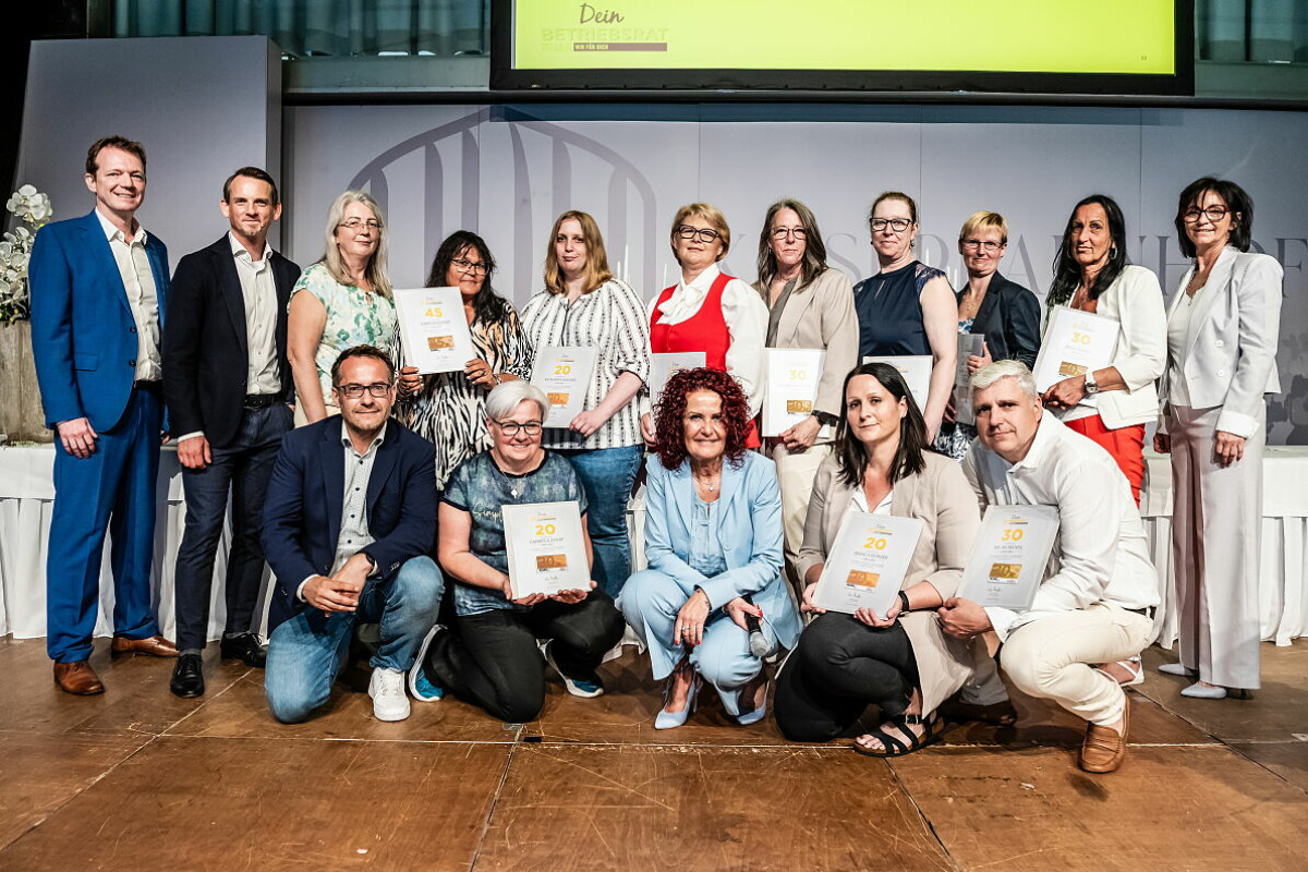 A group of people posing together on a stage, some holding certificates. The background features a white screen with yellow at the top. They are dressed in business or casual attire, and the mood appears celebratory.