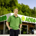 A person in a green polo shirt stands confidently in front of a supermarket with a green "ADEG" sign. There are shopping carts in the background, and trees are visible behind the building.