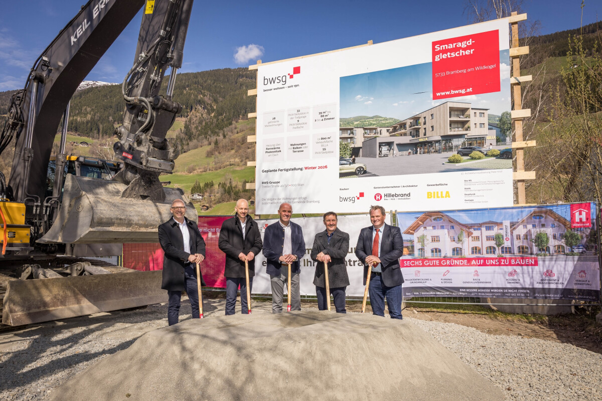 Five people in formal attire stand with shovels on a mound of dirt at a construction site. Behind them is an excavator and a large billboard displaying the new building project. The background features scenic hills and trees.
