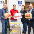 Four people standing outside a building, holding paper bags. One person holds a sign that says "Danke!" The entrance displays a sign for "Caritas & Du." A box with a donation-themed design is in the foreground.
