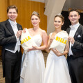 Two men and two women dressed in formal attire, each holding a bag labeled "BILLA," standing in a wood-paneled room. The women are wearing white dresses and tiaras, while the men are in black tuxedos.