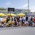 A large group of children and adults posing on an outdoor basketball court. Some are holding basketballs, and there is a basketball hoop in the background. Yellow umbrellas and a police vehicle are visible behind them.