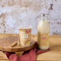 A jar of oat yogurt and a bottle of oat milk on a wooden surface with a rustic background. A brown cloth and an ear of wheat are placed beside them.