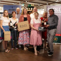 A group of people smiling and holding wine bottles and boxes in a wine shop. They are standing on a tiled floor, dressed in casual and traditional attire. There are wine bottles displayed in the background.
