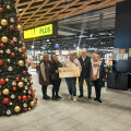 Five people standing in a supermarket near a large Christmas tree holding a large check. The supermarket has a Billa Plus sign in the background. The group is smiling and dressed in winter clothing.