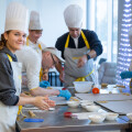 People wearing aprons and chef hats are gathered around a table participating in a cooking class. They are preparing ingredients with utensils and bowls, and one woman in the foreground is smiling at the camera. The setting appears bright and modern.