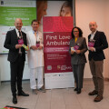 Four people standing in front of a banner that reads "First Love Ambulanz," holding informational brochures. Two are in suits, one in a white medical coat, and one in business attire. The setting appears to be a medical or health facility. The floor has a line with "Ambulanz" written on it.