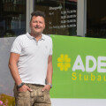A man in a white polo shirt standing outside a store with a sign that reads "ADEG Stubauer." There are flowers in the foreground, and business hours are displayed on the glass window behind him.