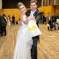 A couple in formal attire, with the woman in a white gown and tiara, and the man in a black suit, smiling as they dance in a large hall. They are holding a bag with the word "JILLA" visible. Other people in formal wear are in the background.
