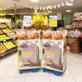 Grocery store entrance with fruit and vegetable displays on the left and shelves with packaged goods on the right. Two large stands with shopping bags featuring the text "Einfach Helfen" are in the center. There are baskets of bread on the right and a decorated ceiling banner.