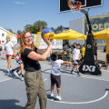 A woman smiling and holding a basketball on an outdoor court, with children playing basketball in the background on a sunny day.