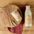 A carton of bio oat yogurt and a glass bottle of milk placed on a round wooden board with wheat stalks, all set on a wooden table with a red cloth beneath.