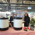 Two people standing behind a table with a red and white checkered tablecloth, featuring two large white containers. A small decorated Christmas tree is on the table. They are inside a store with shelves and products in the background.