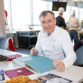 A chef in a white uniform sits at a table, signing a large book. The table is filled with various pastries and magazines. Several chefs in white hats are working in the background. The room has large windows and festive decor.