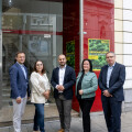 Five people standing in front of a storefront with the signs "Billa" and "Regionalbörse" visible. They are dressed in business casual attire, and the background includes a glass entrance and partial view of a building facade.