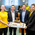 Four people stand in a room holding a large decorated cake. Two individuals wear yellow shirts, while the other two are in suits. Seated participants in yellow shirts are visible in the background.