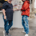 Two men standing outside a food stand, smiling at the camera. One wears a dark apron and cap, the other a red jacket. They stand next to a sign featuring an illustration and text. The location appears to be urban with columns in the background.